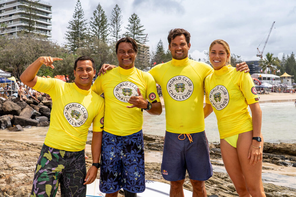 eam Tahiti surfers smiling together on the beach at the World Club Championship Surfing, wearing bright yellow competition rash vests with the event logo.