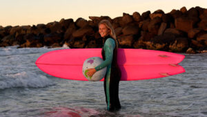 Steph Gilmore standing in the water with a bright pink surfboard and globe, smiling at sunrise, supporting the World Surfing Conservation Conference 2026 on the Gold Coast.